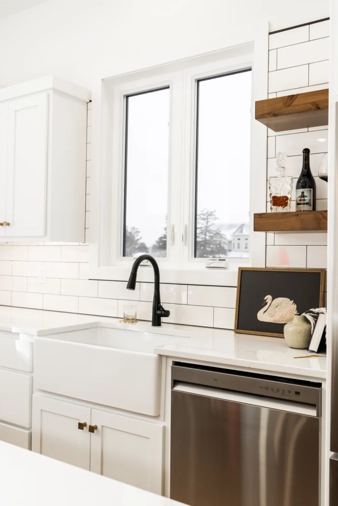 Modern kitchen interior featuring white cabinetry, farmhouse sink, black faucet, open shelving with decorative items, and large windows, showcasing a bright and inviting atmosphere ideal for custom home design.