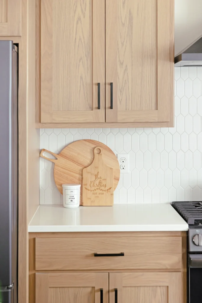 Modern kitchen design featuring light wood cabinetry, a white hexagonal backsplash, a wooden cutting board with engraved text, and a candle on a white countertop, emphasizing custom home aesthetics.