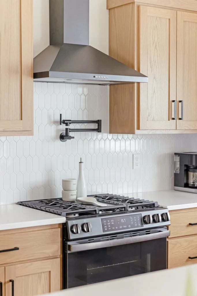 Modern kitchen featuring a stainless steel gas range, sleek range hood, and light wood cabinetry against a white hexagonal tile backsplash, showcasing a stylish and functional cooking space ideal for custom home design.